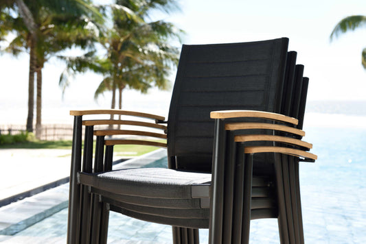 Dark gray stacking armchairs with teak accents beside a pool, palm trees visible in background