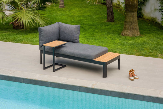 A modern outdoor furniture set with a black metal frame and dark grey cushioned seat. A side table is attached, positioned near a swimming pool backdrop with green grass and trees.