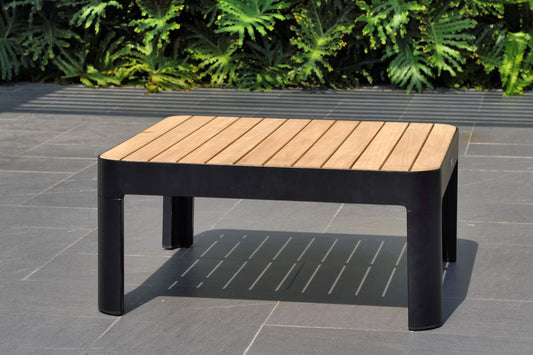 A wooden dark square coffee table with a light brown tabletop and black legs, placed outdoors on a tiled floor with greenery in the background.