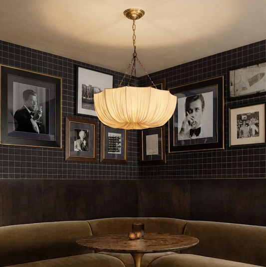 A Scallop-edged fabric hanging lamp with a brass fixture, hanging from the ceiling in a room with black tiled walls and framed black and white photos arranged on the wall.