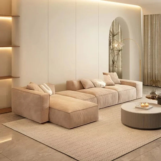 Beige sectional sofa set with matching ottoman and cushions, set in a modern living room with minimalist decor, neutral walls, and round wooden side table.