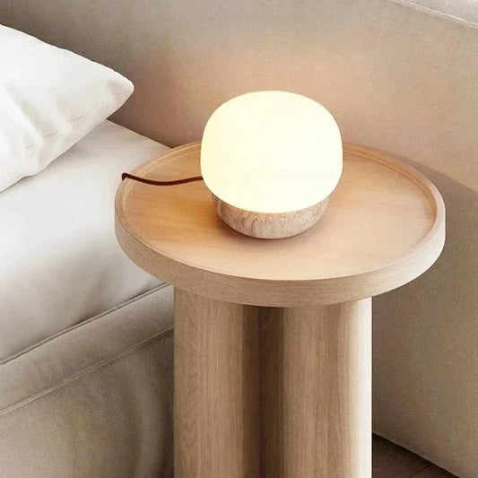A mushroom-shaped lamp on a round wooden bedside table with a white wall and a bed in the background.