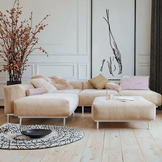 Beige velvet corner sofa set with white metal legs, adorned with soft throw pillows in muted hues, paired with a circular rug and minimalistic decor in a light-toned wooden space.
