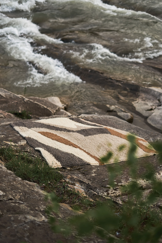 Hand woven area rug 'Stone' on rocky terrain next to a flowing stream, beige, brown, and gray stripes