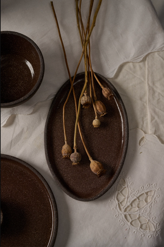 Dark brown oval stoneware serving platter with dried poppy pods, alongside small bowls on a white lace tablecloth.