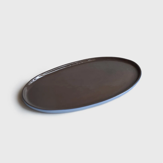 The image shows a smooth, oval-shaped porcelain serving platter with a matte finish. The platter's upper surface is dark grey, and the rim is a light blue color. The background is plain white.