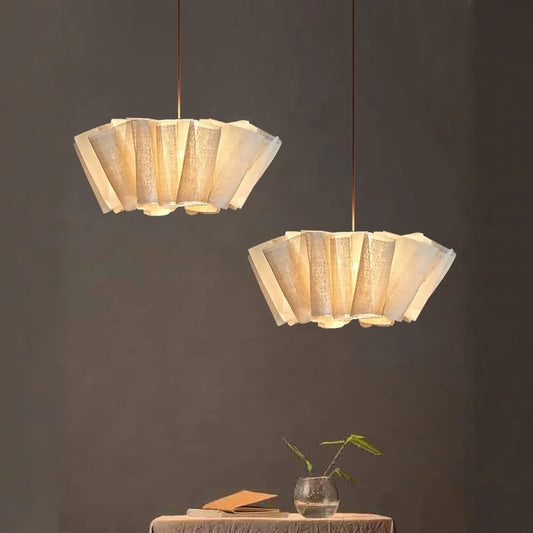 Two Wabi Sabi pleated pendant lights with Nordic linen fabric, hanging from dark wires against a neutral background; one table with a plant and book beneath them.