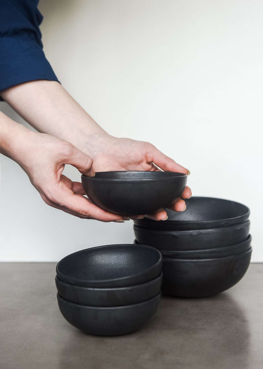 Hand holding a matte black stoneware sauce bowl with multiple bowls stacked on a concrete surface.