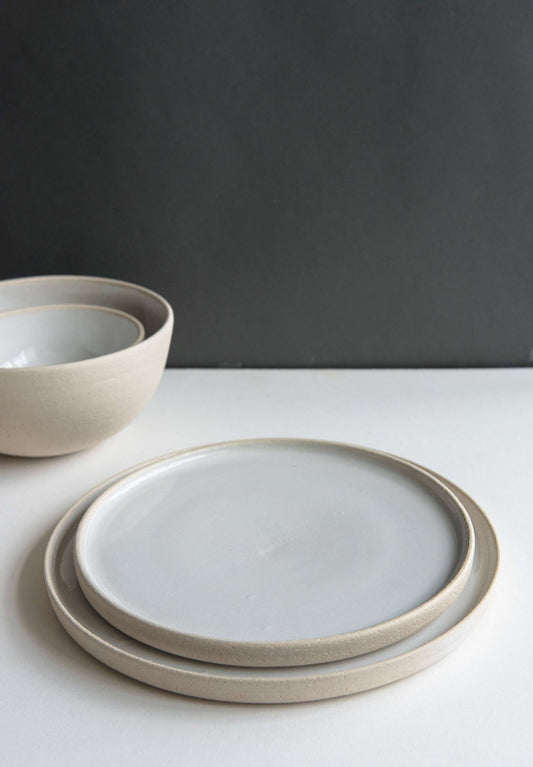 Stacked handmade stoneware plates with a light grey matte finish and off-white rim, placed on a white surface against a dark background.