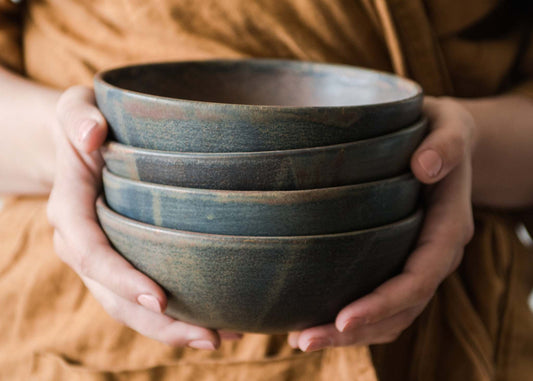 Hands holding a stack of dark brown rustic stoneware soup bowls on a wooden surface.