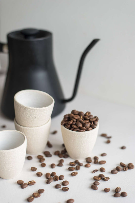 White stoneware espressos cups with coffee beans beside a black pour-over kettle on a white surface;