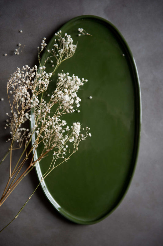 Handmade oval porcelain platter in green, placed on dark surface with white dried flowers on the left.