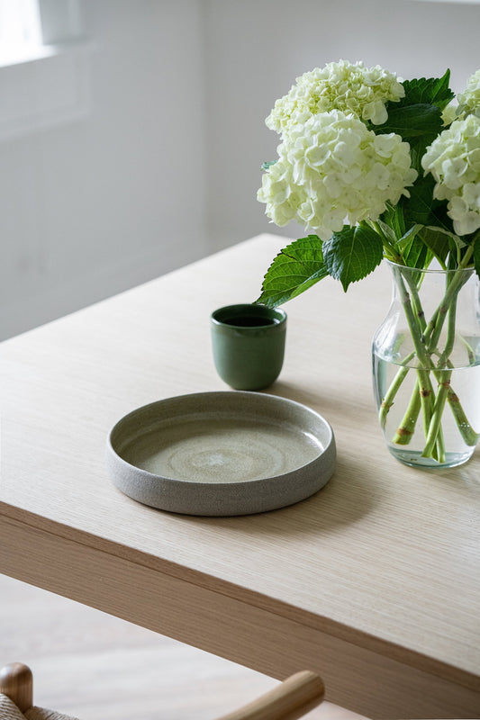 Handmade Stoneware Dinner Plate on wooden table, surrounded by hydrangeas in vase and green mug, indoor setting.