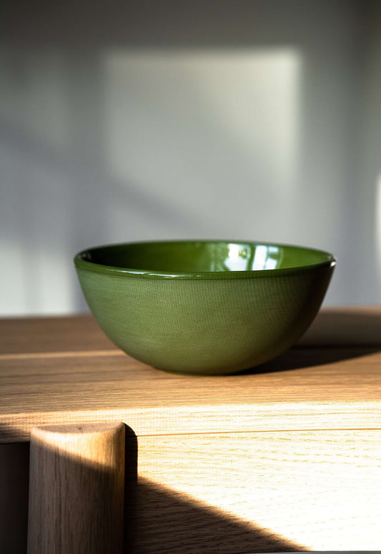 A green handmade porcelain bowl坐 on a wooden surface with warm sunlight casting shadows.