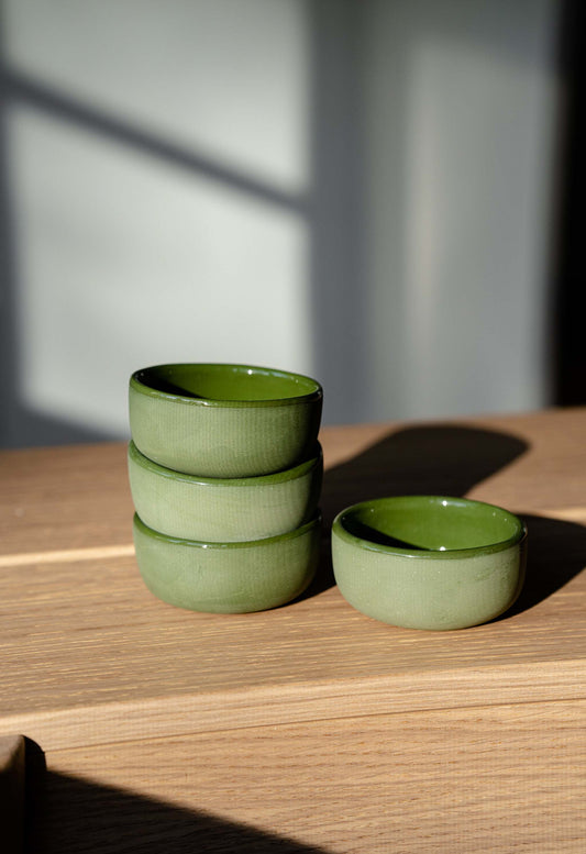 Handmade porcelain sauce bowls with a soft green hue on a wooden surface with natural light casting shadows.