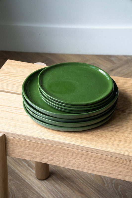 Stack of six green handmade porcelain dinner plates on a wooden table against a plain wall.