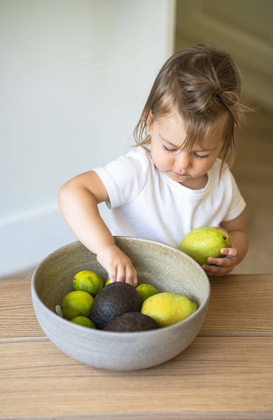 A toddler arranging avocados in a large stoneware bowl on a wooden table.