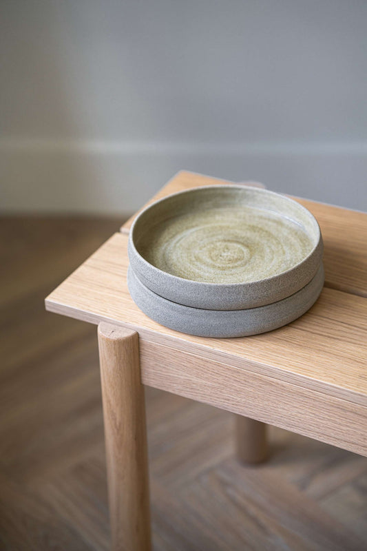 Stack of three stoneware salad plates with high sides, light grey in color, displayed on a wooden stool against a neutral background.