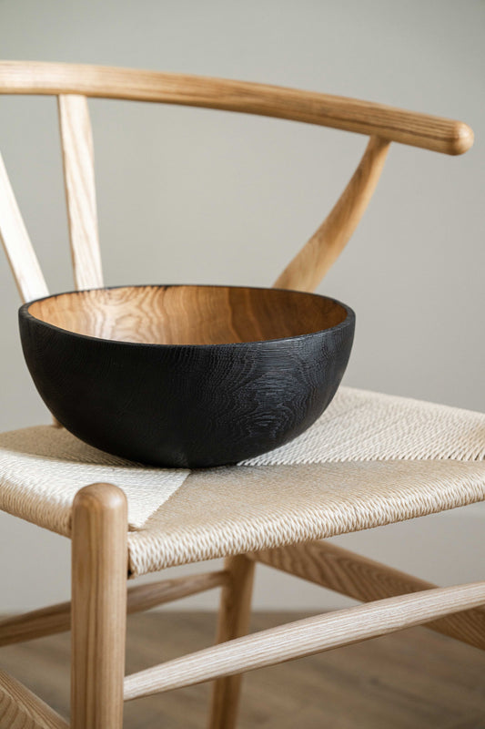 A black hand-carved wooden bowl with a charred finish sits on a wooden chair with a woven seat against a plain background.
