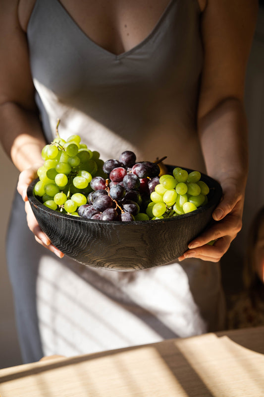 Wooden bowl filled with green and purple grapes, held by person wearing a dress