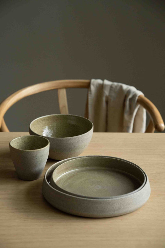 Handmade stoneware dinner set with concrete texture. Olive green color. Setup on wooden table with chair in background.