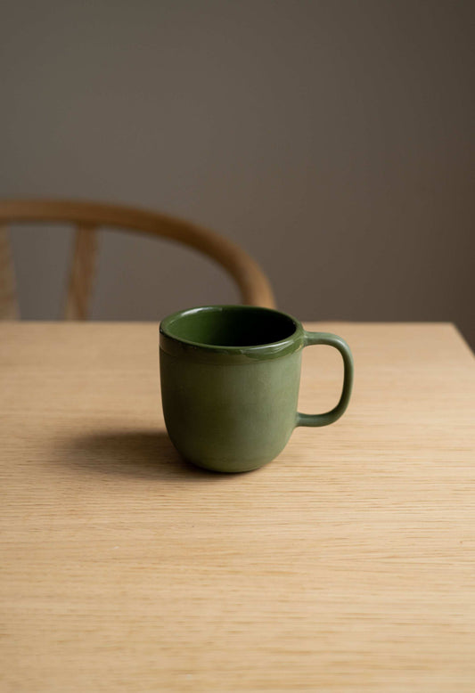 A green, glazed porcelain coffee mug on a wooden table with a chair in the background.