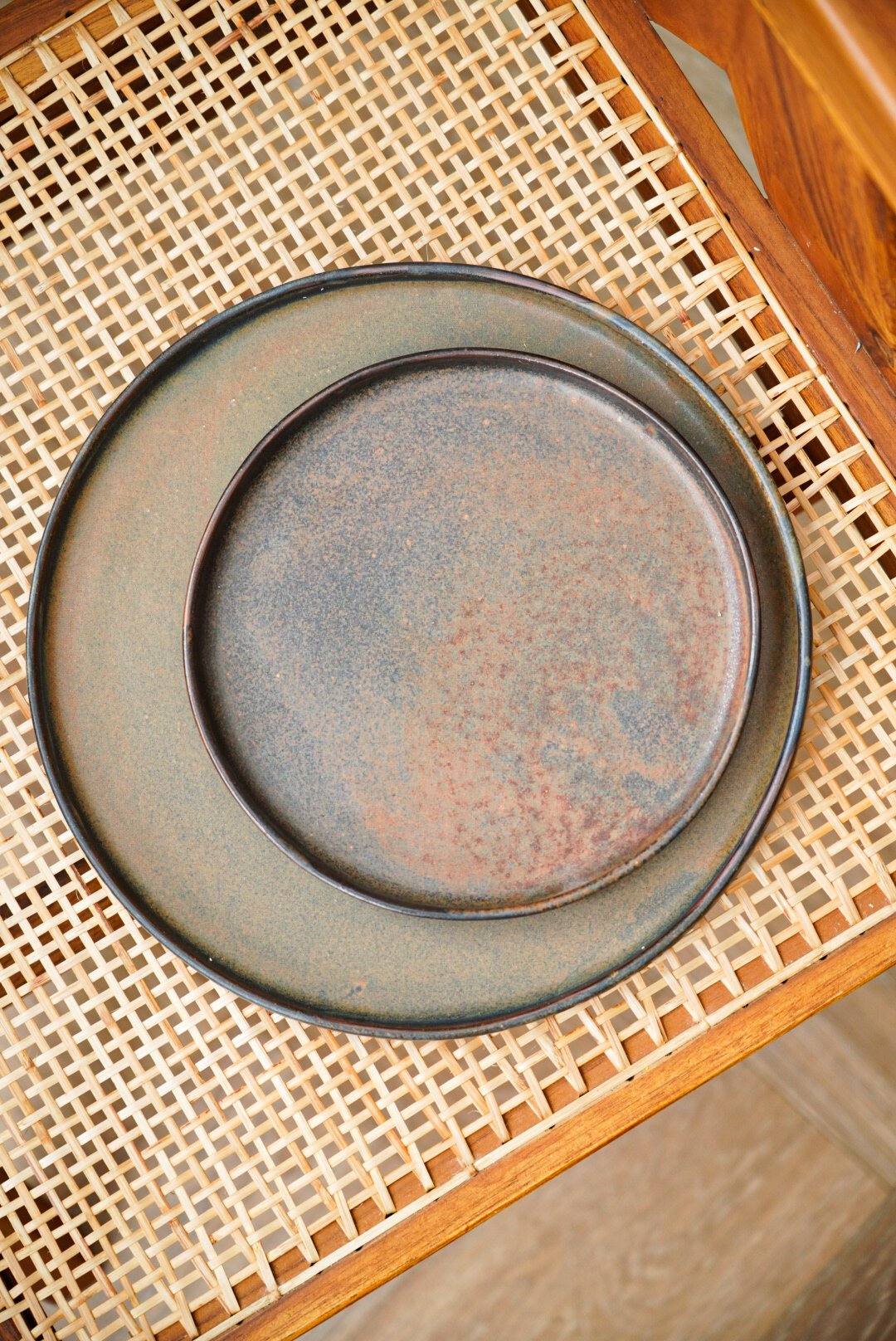 Rustic dinner plate with a matte brown finish on a woven wicker tray, placed on a wooden table.