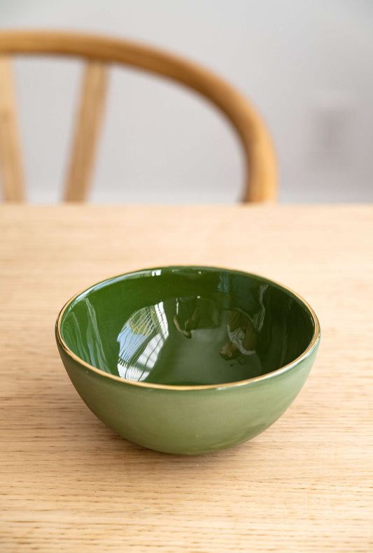 Green handmade porcelain bowl with gold rim on wooden table, chair in background