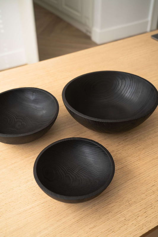 Three black charred wood salad bowls on a wooden surface, showcasing their natural grain patterns.
