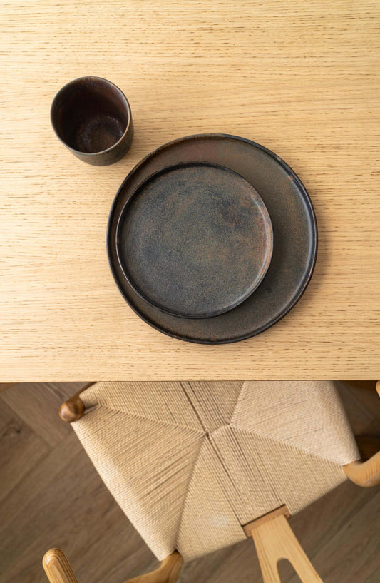 Dark stoneware plates with textured finish on a wooden table with woven chair seat