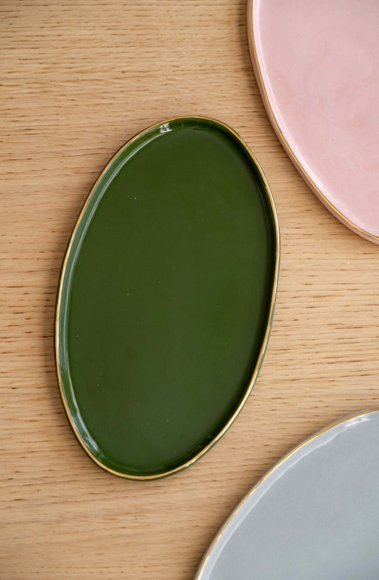 A green porcelain oval serving platter with a gold rim, placed on a wooden surface next to a pink plate and another platter.