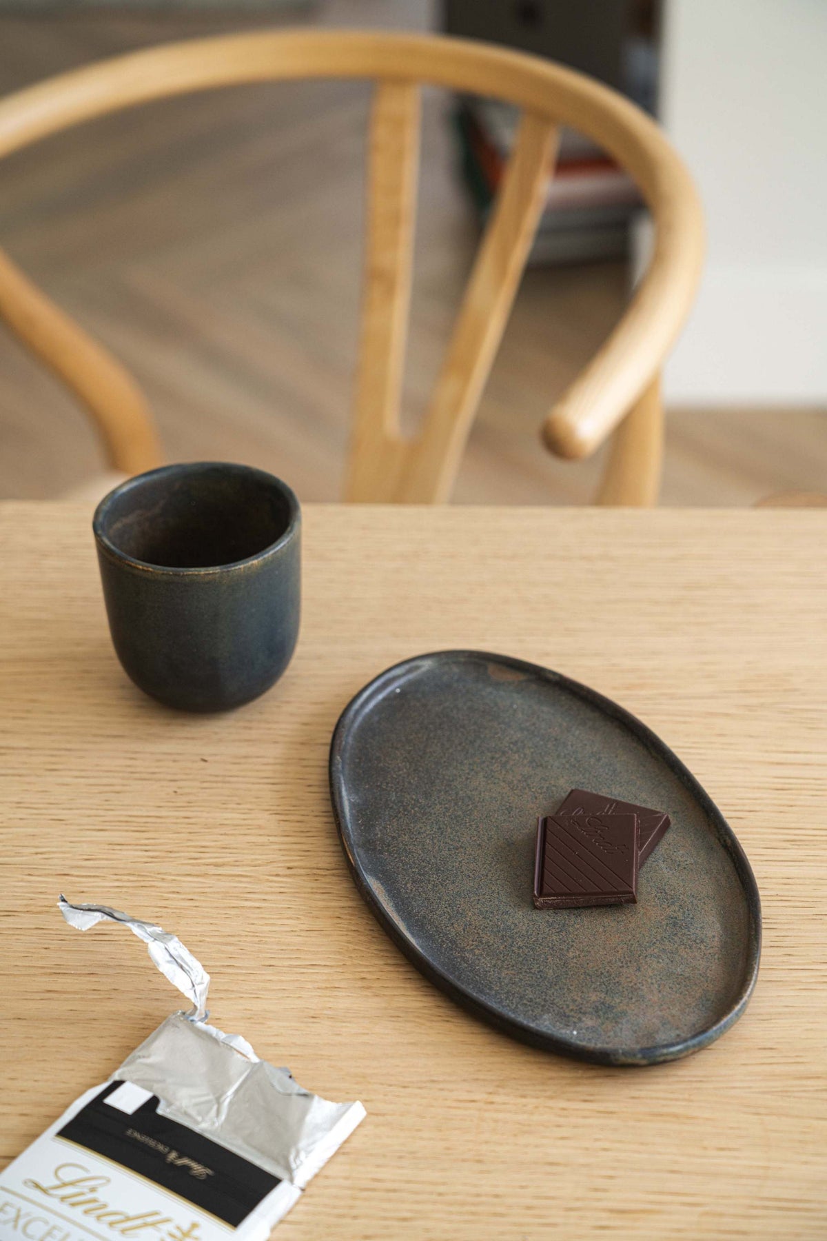Rustic matte stone serving platter with dark chocolate on a wooden table, minimalist interior with wooden chair, Lindt chocolate wrapper.