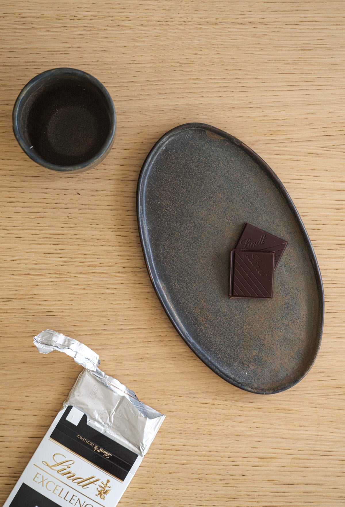A grey oval serving platter on a wooden surface with two Lindt chocolates and a dark-rimmed bowl beside it.