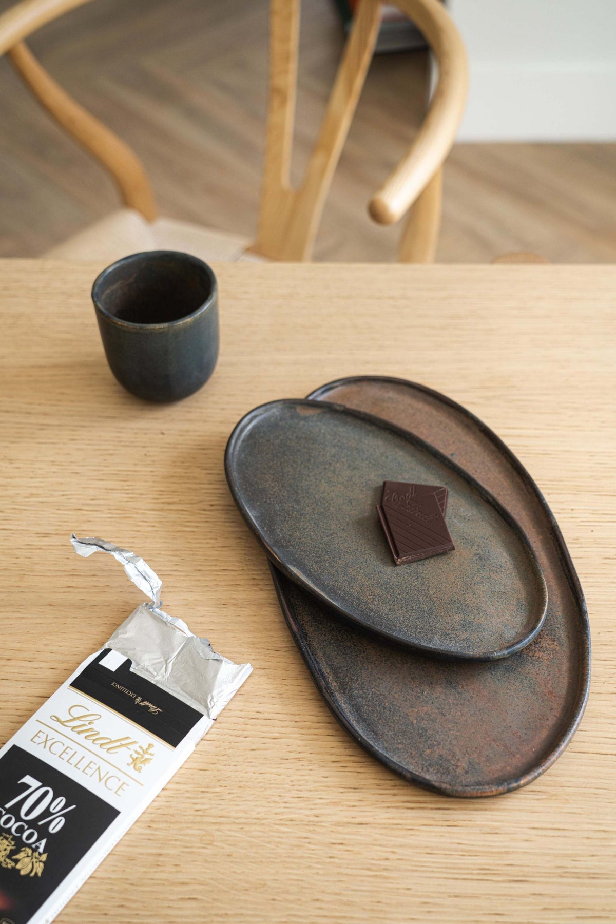 Rust Matte Stoneware Serving Platter on a wooden table with a Lindt Excellence chocolate bar and a black ceramic drink cup, soft background with a wooden chair.