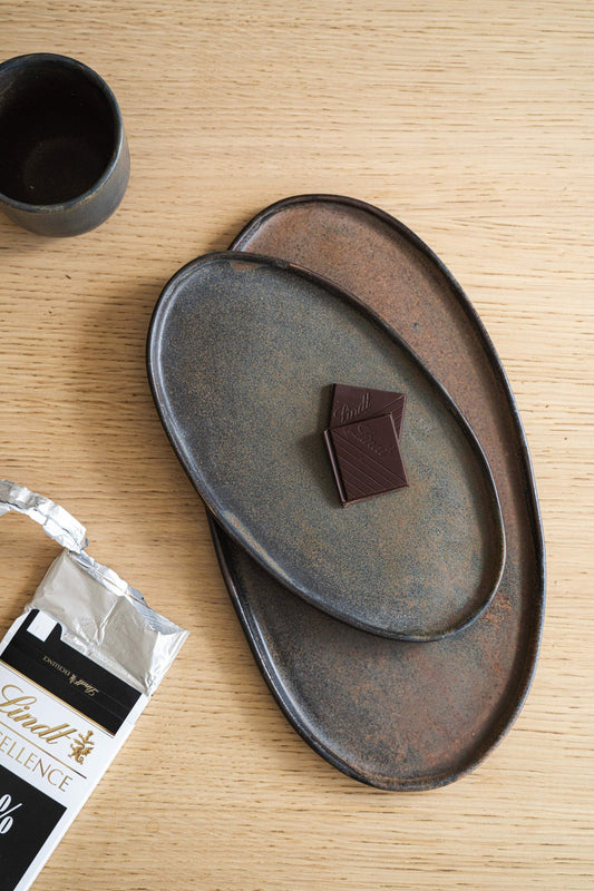 A set of two matte stoneware serving platters with a matte finish, in a dark rust color. One platter holds a piece of Lindt chocolate on a textured wooden surface. Nearby is a dark cup and Lindt chocolate packaging.