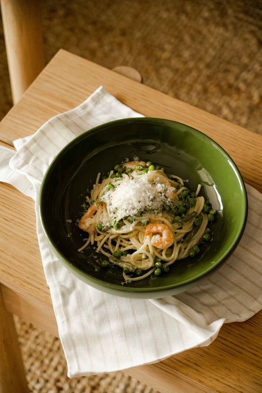 Green bowl with pasta, peas, shrimp, and parmesan cheese on a wooden table with a white cloth.