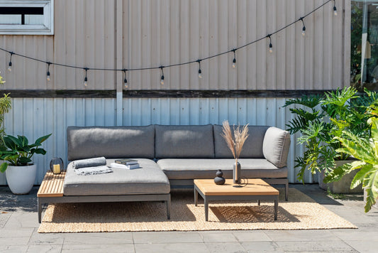 Outdoor sectional with gray cushions and wooden legs, set up on a patio with plants and a string of lights.