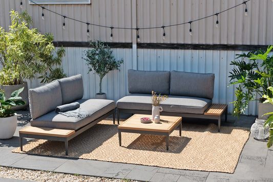 Gray sectional sofa and lounge with gray cushion and wooden tray tables on woven rug outdoors with potted plants and string lights.