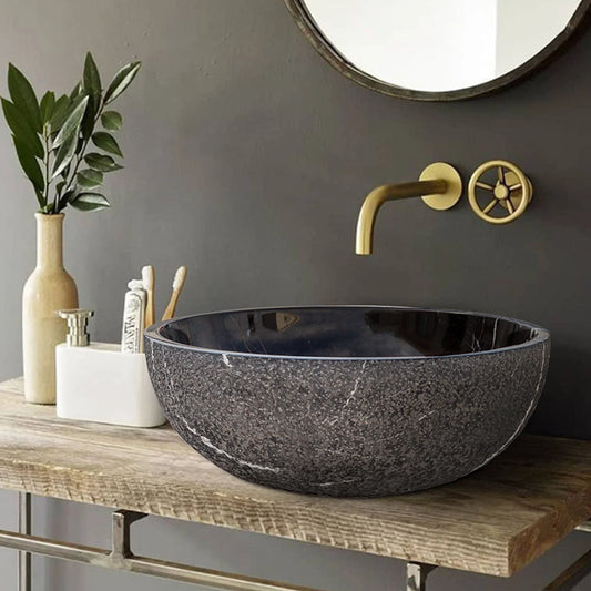 A black marble vessel sink 16 inches round with a polished interior and rough exterior, set in a rustic bathroom counter with gold faucet and greenery.