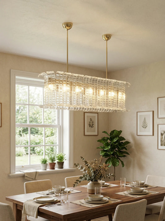 Rectangular Murano glass chandelier with clear beads, hanging above a dining table with plates and glasses, natural light through a window with small plants on the sill, and framed botanical prints on the wall.