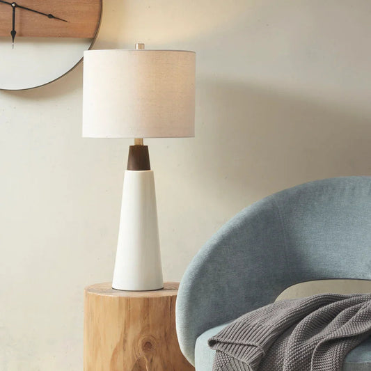 Conical ceramic table lamp with two-toned finish and off-white drum shade, placed on wooden side table in mid-century modern setting.