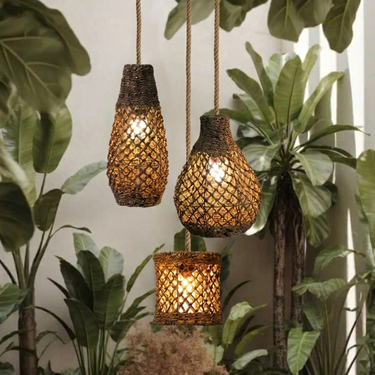Three woven jute pendant lights hanging, with a warm glow from the bulbs, positioned against a backdrop of lush greenery, likely in an indoor setting.