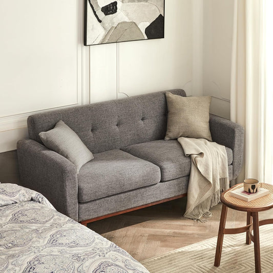 A mid-century modern gray loveseat sofa with tufted backrest, wooden legs, beige throw, white blanket, patterned bedspread, and abstract artwork in a minimalist setting.