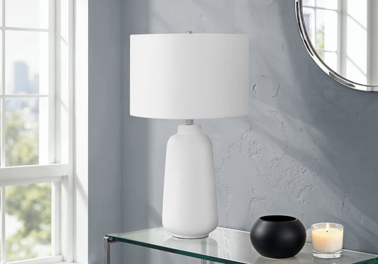 Modern white textured table lamp with white shade on a glass table, grey wall backdrop, and natural light from window, black bowl beside it, white candle in glass holder, round mirror in background