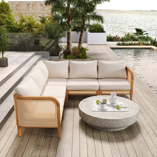 An acacia wood sofa with white cushions on a wooden deck by a water view, featuring a round concrete coffee table with white tableware and fruit.
