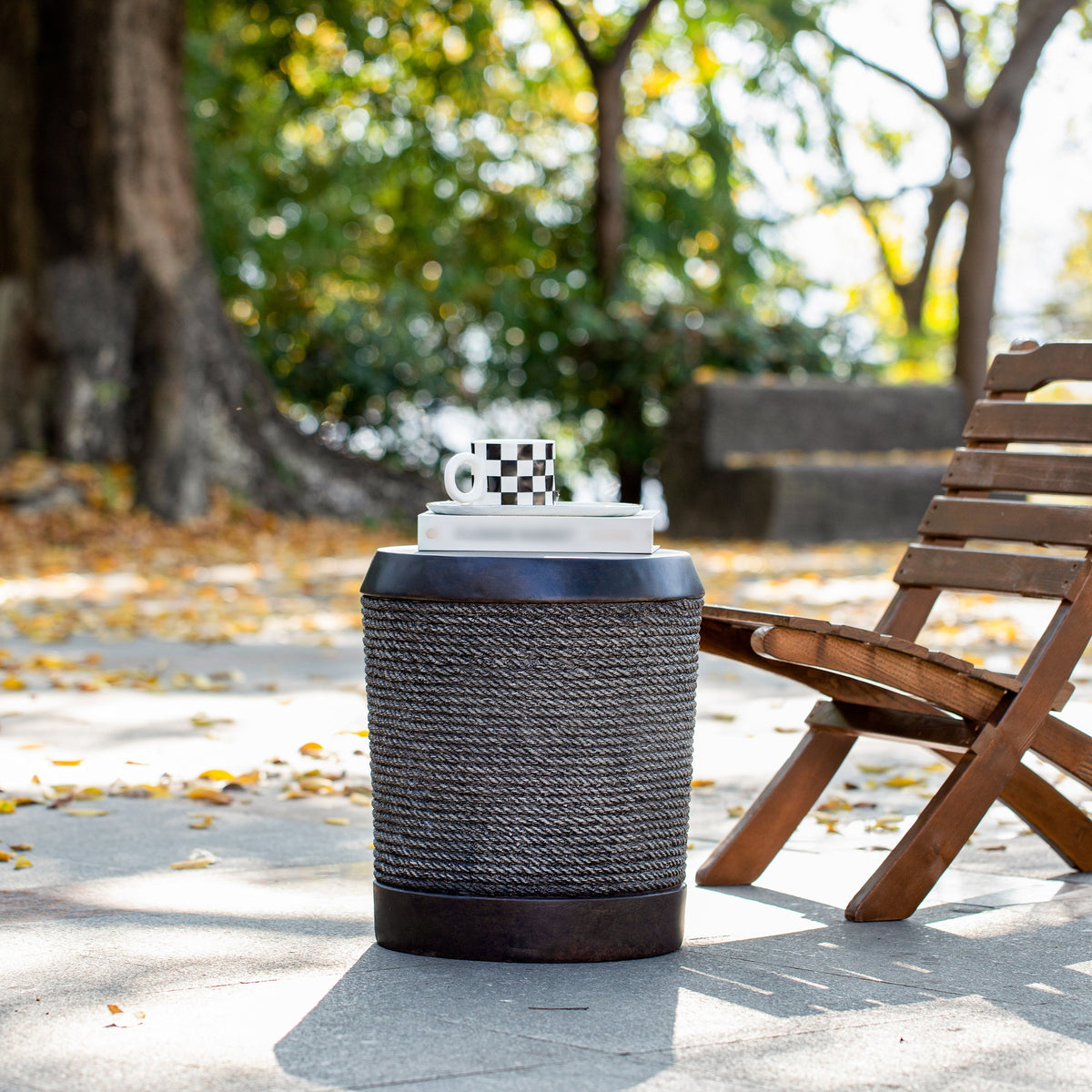Outdoor table with braided cord texture, dark brown color, and vintage trim, placed on a paved path near a wooden bench and a tree.