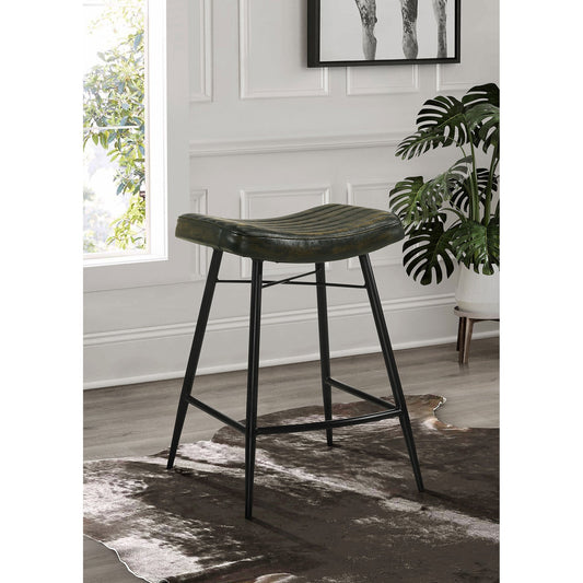 A green leather bar stool with black metal legs in a room with a wooden floor, wall, and window. A monstera plant is in the background.