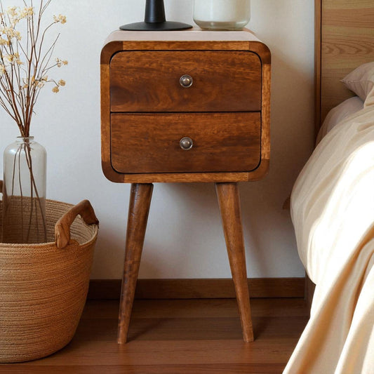 A Soli Wood Mini Chestnut Curved Nightstand with a natural walnut finish, two drawers, and a silver knob, placed in a bedroom next to a bed, a basket, and a vase with dried flowers.