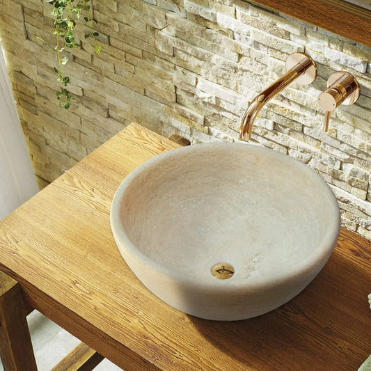 A beige travertine vessel sink with honed finish on a wooden table, set against a stone wall.