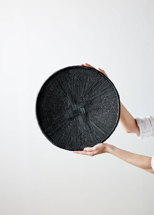 Hands holding a circular, hand-painted black Binga flat basket against a plain background.